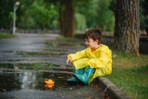 criança jogando com brinquedo barco dentro poça. criança jogar ao ar livre de chuva. outono chuvoso clima ao ar livre atividade para jovem crianças. criança pulando dentro turvar poças. à prova d'água Jaqueta e chuteiras para bebê. infância foto