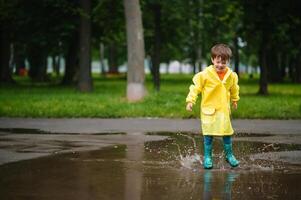 pequeno Garoto jogando dentro chuvoso verão parque. criança com guarda-chuva, à prova d'água casaco e chuteiras pulando dentro poça e lama dentro a chuva. criança caminhando dentro verão chuva ao ar livre Diversão de qualquer clima. feliz infância. foto