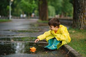 criança jogando com brinquedo barco dentro poça. criança jogar ao ar livre de chuva. outono chuvoso clima ao ar livre atividade para jovem crianças. criança pulando dentro turvar poças. à prova d'água Jaqueta e chuteiras para bebê. infância foto