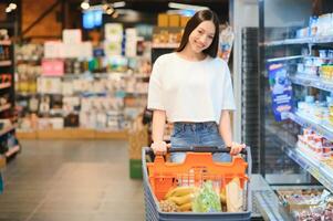 jovem satisfeito Diversão mulher dentro casual roupas compras às supermercado loja com mercearia carrinho foto