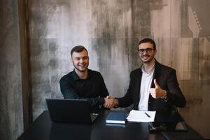 dois empresários dentro a escritório sorridente às a Câmera enquanto trabalhando juntos atrás uma computador portátil computador. foto