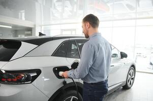 barba homem tentando uma Novo cobrando cabo com uma carro cobrando estação às a motor concessionária. conceito do comprando elétrico veículo. inteligente ecológico vivo foto