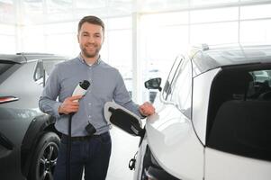 barba homem tentando uma Novo cobrando cabo com uma carro cobrando estação às a motor concessionária. conceito do comprando elétrico veículo. inteligente ecológico vivo foto