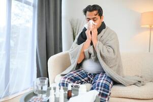 doente homem sentado às casa em sofá dentro vivo sala, espirros tem alergia e escorrendo nariz, indiano homem com guardanapo tem uma frio foto