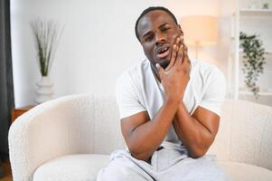 fechar-se foto do uma jovem afro-americano homem sofrimento a partir de uma forte dor de dente às casa em a sofá. ele detém dele mão para dele bochecha, fazendo careta dentro dor