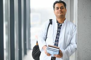 retrato do uma jovem indiano masculino médico aluna dentro uma branco casaco acenando foto