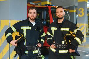 retrato do dois jovem bombeiros dentro uniforme em pé dentro a fogo estação foto