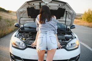 atraente fino jovem menina dentro verão calção e camisa reparos uma quebrado carro. uma lindo mulher carrinhos perto elevado carro capuz. foto