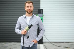 bonito barbudo homem dentro casual vestir, em pé às a cobrando estação e carregador para a elétrico carro. eco elétrico carro conceito foto