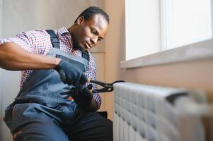 afro reparador dentro macacão usando Ferramentas enquanto instalando ou reparação aquecimento radiador foto