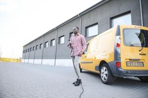 africano homem segurando carregar cabo dentro em mão em pé perto elétrico carro. foto