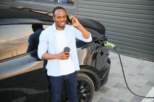 africano homem em pé perto elétrico carro com cobrando cabo dentro plugue. eco amigáveis veículo cobrando em estação. estilos de vida conceito foto