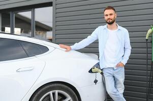 em pé e esperando para carregar para terminar. jovem à moda homem é com elétrico carro às dia foto