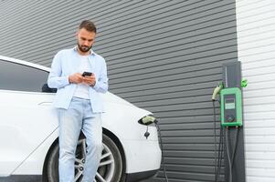 casual homem com Smartphone perto elétrico carro esperando para a terminar do a bateria cobrando processo foto