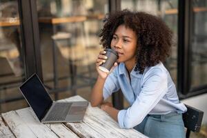 jovem afro-americano mulher segurando café copo sentado lado de fora cafeteria com computador portátil. elevado uma copo do café e bebido e encarou longe foto