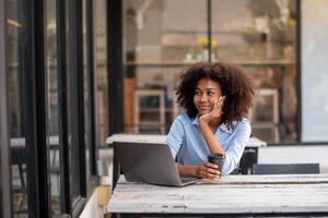 lindo jovem africano mulher segurando café copo sentado lado de fora cafeteria com computador portátil juntou com outro, interessado dentro o que é em volta, dentro a humor do felicidade foto