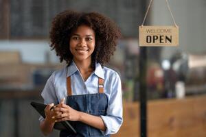 fêmea africano café fazer compras pequeno o negócio proprietário vestindo avental em pé e dentro a fundo lá é uma bem-vinda placa foto