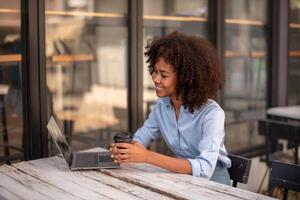 Preto mulher interessado dentro alguma coisa em computador tela computador conversando, segurando copo do café dentro mãos socializar com amigo foto