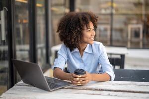 jovem afro-americano mulher segurando café copo sentado lado de fora cafeteria com computador portátil. Veja Fora para interessante coisas e sorrir às seu arredores. foto