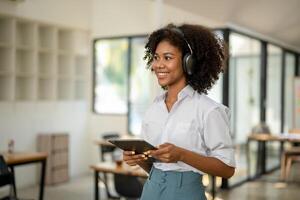africano americano Faculdade aluna com fones de ouvido dentro mão com tábua em pé sorridente olhando frente dentro cafeteria ou área de trabalho foto