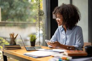 africano americano mulher sentado dentro frente do uma computador portátil e segurando uma Móvel telefone foto