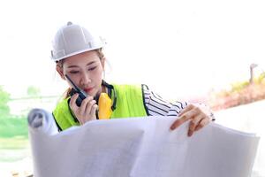 ásia fêmea engenheiro segurando plantas e rádios coordenada com a construção equipe às a ao ar livre local. conceito do construção construção, real Estado desenvolvimento. foto