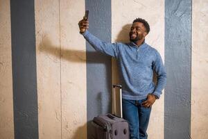 feliz homem com mala de viagem levando selfie com uma telefone enquanto inclinado contra uma parede dentro passagem do a estrada de ferro estação. cópia de espaço em imagem para seu advertir ou texto. foto