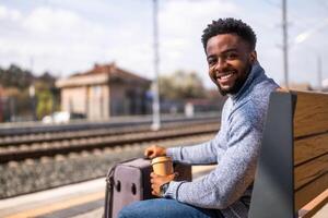 feliz homem com mala de viagem goza bebendo café enquanto sentado em uma Banco às a estrada de ferro estação. foto