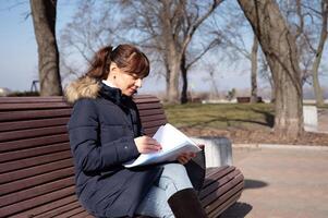 uma jovem fêmea contador senta em uma parque Banco e parece através comunicando documentos durante dela pausa foto