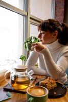 uma jovem mulher sentado dentro uma cafeteria e bebendo chá, olhando Fora a janela e esperando para dela parceiro foto