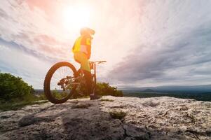 uma jovem cara senta em uma montanha bicicleta dentro uma montanhoso área contra a pano de fundo do uma privado setor. a Sol é brilhando sobre a ciclista foto