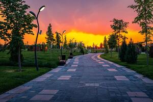 cidade parque dentro cedo verão ou Primavera com pavimento, lanternas, jovem verde grama, árvores e dramático nublado céu em uma pôr do sol ou nascer do sol. foto