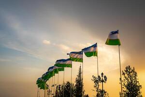 bandeiras do uzbequistão acenando em uma pôr do sol ou nascer do sol dramático céu fundo. foto