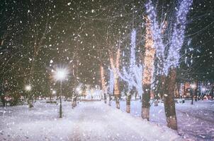queda de neve dentro uma inverno parque às noite com decorações, brilhando lanternas, calçada coberto com neve e árvores vintage filme estética. foto