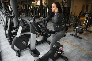 jovem mulher com fones de ouvido fazendo exercícios em papelaria bicicleta dentro uma Academia ou ginástica Centro. jovem desportivo mulher dentro Academia ouço música a partir de Smartphone. mulheres fazendo cardio exercícios foto