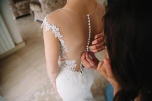 dama de honra preparando noiva para Casamento dia. dama de honra ajudando noiva apertar laço dela Casamento branco vestir antes cerimônia. luxo nupcial vestir fechar acima. Casamento manhã momentos detalhes conceito. foto