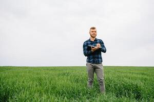 jovem agrônomo detém caderno dentro verde trigo campo. agronegócio conceito. agrícola engenheiro em pé dentro uma trigo campo com uma tábua dentro verão. foto