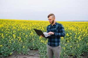 jovem agrônomo detém computador portátil dentro estupro campo. agronegócio conceito. agrícola engenheiro em pé dentro uma estupro campo com uma computador portátil dentro verão. foto