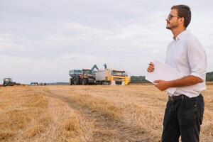 jovem atraente agricultor com computador portátil em pé dentro trigo campo com combinar colheitadeira dentro fundo. foto