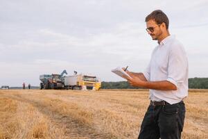 feliz jovem agricultor engenheiro com caderno em pé em trigo campo enquanto combinar colheitadeira trabalhando dentro fundo. foto