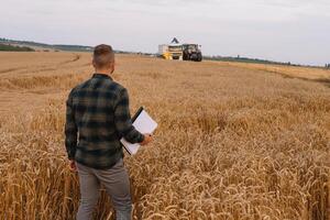 jovem atraente agricultor com computador portátil em pé dentro trigo campo com combinar colheitadeira dentro fundo. foto