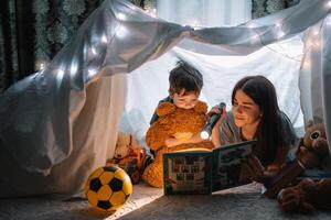 feliz família mãe e criança filho lendo uma livro com uma lanterna dentro uma barraca às lar. família conceito. foto