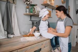 jovem feliz mãe e dela bebê cozinhar biscoitos às casa dentro a cozinha. Natal caseiro Pão de gengibre. fofa Garoto com mãe dentro branco uniforme e chapéu cozinhou chocolate biscoitos foto