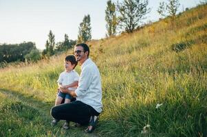 bonito Papai com dele pequeno fofa filho estão tendo Diversão e jogando em verde gramíneo grama. feliz família conceito. beleza natureza cena com família ao ar livre estilo de vida. família em repouso junto. pais dia. foto