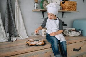 jovem feliz mãe e dela bebê cozinhar biscoitos às casa dentro a cozinha. Natal caseiro Pão de gengibre. fofa Garoto com mãe dentro branco uniforme e chapéu cozinhou chocolate biscoitos foto