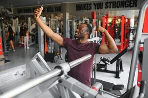 sorridente jovem desportista mostrando bíceps e levando selfie com Smartphone dentro academia. foto