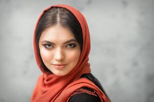 retrato do uma bonita jovem caucasiano mulher com Maquiagem dentro uma vermelho lenço de cabeça em cinzento fundo. foto