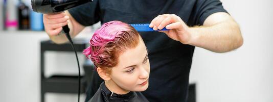 uma cabeleireiro é secagem a Rosa cabelo do a jovem mulher dentro uma beleza salão. foto