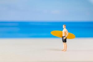 miniatura pessoas homem dentro uma roupa de banho, e segurando uma amarelo prancha de surfe em a de praia foto