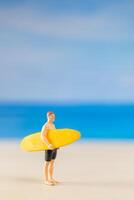 miniatura pessoas homem dentro uma roupa de banho, e segurando uma amarelo prancha de surfe em a de praia foto